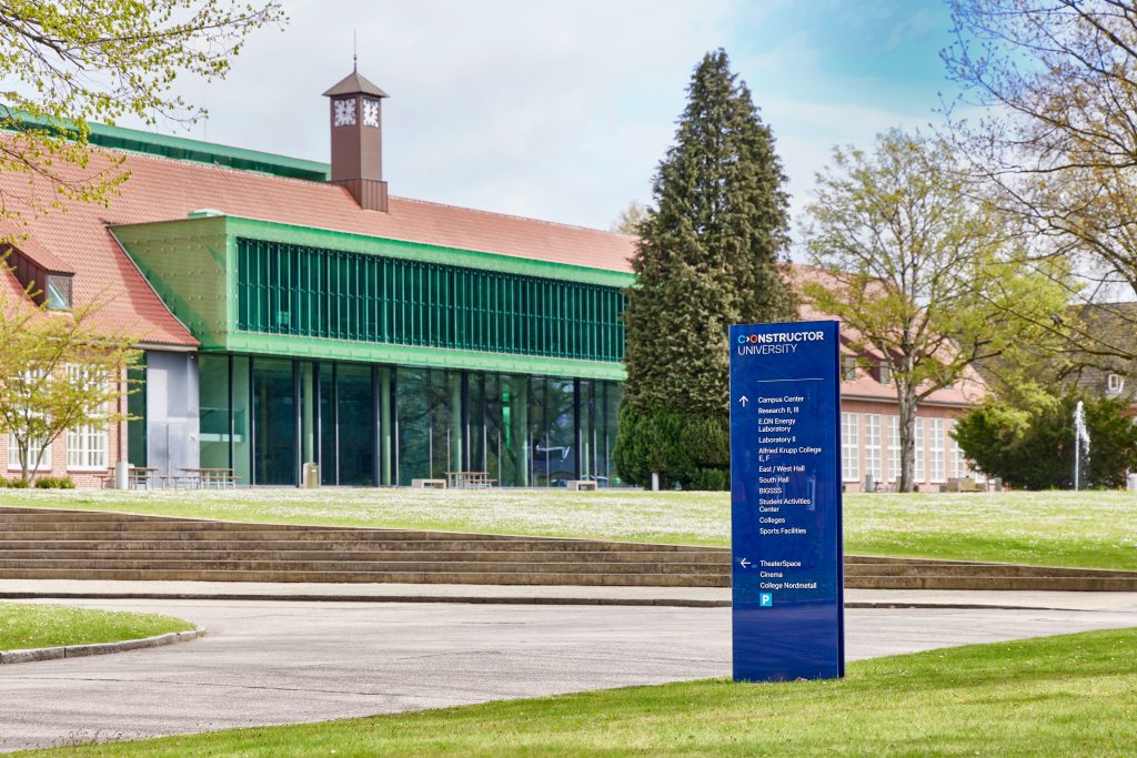 Exterior shot of the IRC with a green-glass entrance, brick walls, and a small clock tower on the roof, and a tall blue campus information sign in front.